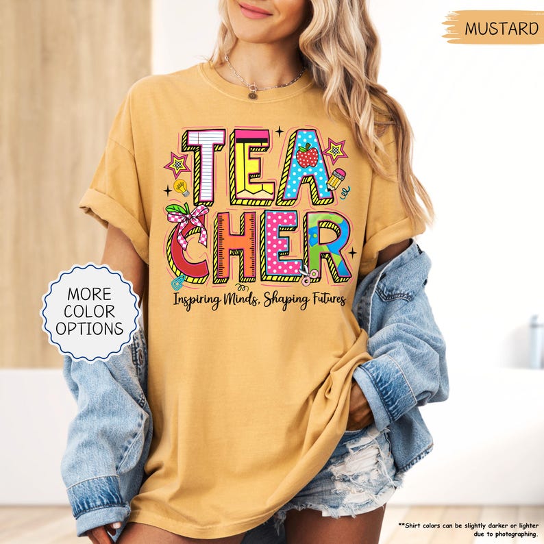 May include: Mustard-coloured t-shirt with the word "TEACHER" in colourful, block letters. The shirt features the phrase "Inspiring Minds, Shaping Futures" below the word "TEACHER". The shirt is a relaxed fit.
