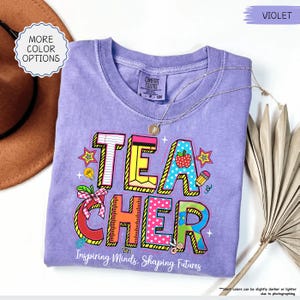 May include: A violet Comfort Colors t-shirt featuring a vibrant "TEACHER" design with an apple, stars, and school supplies. The text "Inspiring Minds, Shaping Futures" is printed below. A gold necklace is draped across the shirt. The shirt is a comfortable fit.
