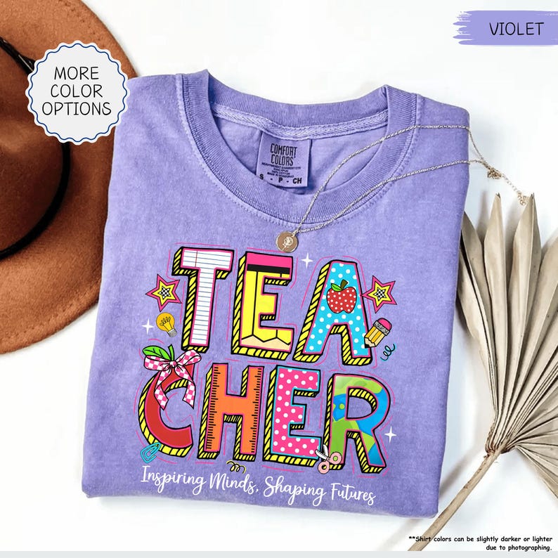 May include: A violet Comfort Colors t-shirt featuring a vibrant "TEACHER" design with an apple, stars, and school supplies. The text "Inspiring Minds, Shaping Futures" is printed below. A gold necklace is draped across the shirt. The shirt is a comfortable fit.