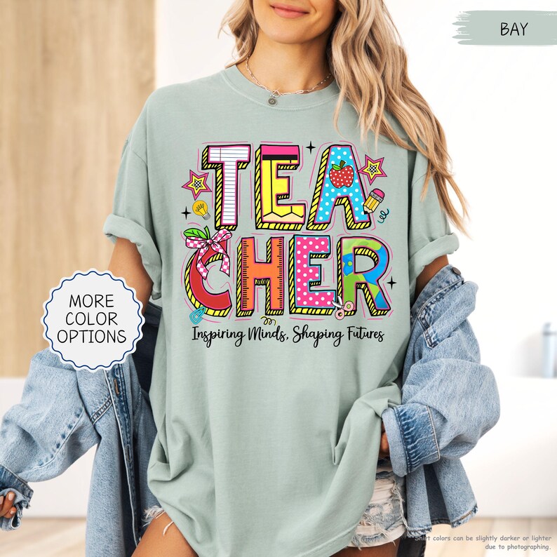 May include: A pale green t-shirt featuring the word "TEACHER" in vibrant, multi-coloured lettering. The design incorporates a strawberry, pencil, and stars. The phrase "Inspiring Minds, Shaping Futures" is printed below.