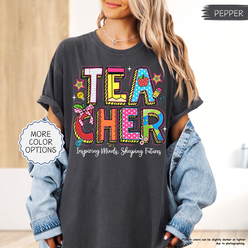 May include: Dark grey t-shirt featuring the word "TEACHER" in colourful, block letters. The design includes stars, an apple, and scissors. The text "Inspiring Minds, Shaping Futures" is below the word "TEACHER".