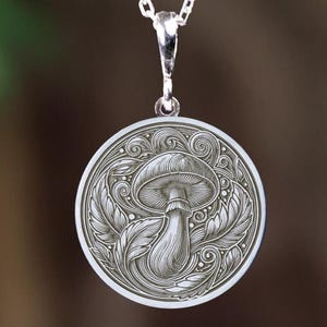 May include: Silver pendant necklace featuring a detailed mushroom design. The circular pendant has an engraved mushroom surrounded by leaves and swirls. The necklace has a delicate silver chain.
