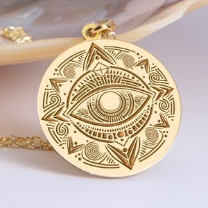 May include: Gold-colored pendant necklace featuring a detailed, engraved design of an eye within a circular frame. The pendant has intricate patterns and is suspended from a delicate gold chain. The necklace is a piece of jewelry.