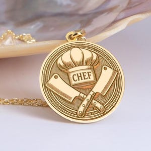 May include: Gold necklace with a round pendant featuring a chef's hat and crossed cleavers. The pendant is engraved with the word "CHEF".