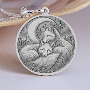 Op de afbeelding: Zilveren ketting met een hanger met een gedetailleerd reliëf van twee vossen onder een maan en sterren. De ronde hanger heeft een gegraveerd ontwerp. De ketting is ook zilver. De diameter van de hanger is ongeveer 2,5 cm.