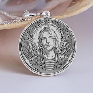 Puede incluir: Collar con colgante de plata con un retrato grabado detallado de un músico con pelo largo, camisa a rayas y chaqueta. Dos guitarras flanquean el retrato, con un diseño de rayos de sol que irradia desde detrás de la cabeza del músico. El colgante es redondo.