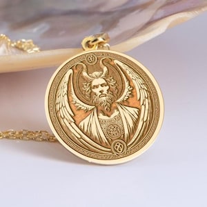 May include: Gold-colored pendant necklace featuring a detailed relief of a horned figure with wings, a beard, and a halo. The pendant is round with an intricate border and is suspended from a delicate gold chain. The design is likely inspired by mythological or religious themes.