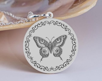 Op de afbeelding: Zilveren ketting met hanger. De ronde hanger heeft een gedetailleerd vlinderontwerp binnen een decoratieve rand. De ketting en sluiting zijn ook zilverkleurig.