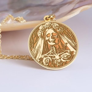 Puede incluir: Un collar de oro con un colgante redondo que presenta una imagen grabada de una novia esqueleto con velo y una corona de rosas.