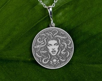 May include: A silver-colored pendant necklace featuring a detailed depiction of Medusa's face surrounded by snakes. The round pendant is suspended from a delicate chain. A skull is at the bottom of the pendant. The necklace is likely made of sterling silver.