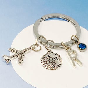 Puede incluir: Llavero plateado con un dije de avión plateado, un dije de globo plateado, un dije de letra R plateada y un dije de piedra preciosa azul.