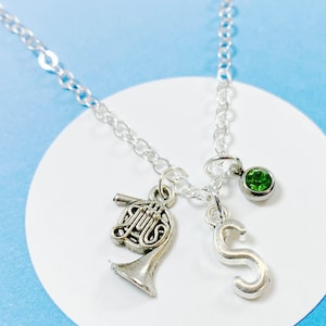 Könnte beinhalten: Eine silberne Halskette mit einem französischen Horn-Anhänger, einem Buchstaben 'S'-Anhänger und einem grünen Edelstein-Anhänger.