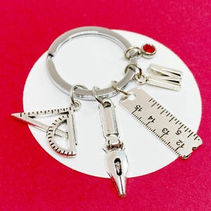 Könnte beinhalten: Silberner Schlüsselanhänger mit einem Lineal, einem Winkelmesser, einer Federkielspitze und einem Anhänger mit dem Buchstaben M. Der Schlüsselanhänger ist an einem silbernen Schlüsselring befestigt.