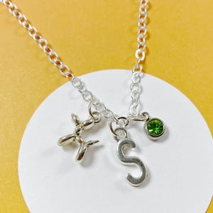 Puede incluir: Un collar de cadena de plata con tres colgantes: un animal de globo, una letra 'S' y una piedra preciosa verde.