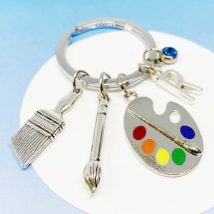 Peut inclure: Porte-clés argenté avec un pinceau, une palette et un charme en forme de lettre 'R'. La palette a des points de peinture colorés et un pinceau dessus.