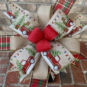 May include: A festive Christmas bow featuring a red truck carrying a Christmas tree on a beige ribbon. The bow has a red center and is accented with plaid and gold trim. The design includes string lights and snow.