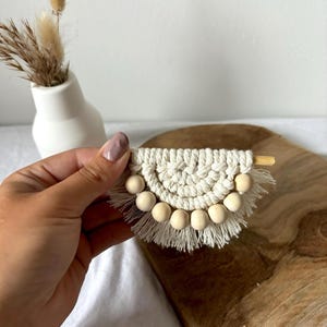 Könnte beinhalten: Ein kleiner, weißer Makramee-Wandbehang in Halbkreisform mit Holzperlen. Der Behang hat unten eine Franse.