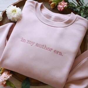 Puede incluir: Una sudadera de cuello redondo en color rosa claro con el texto bordado in my author era. en hilo rosa a juego. La prenda cuenta con cuello acanalado, mangas largas y un forro interior de forro polar suave con un diseño sencillo.