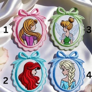 May include: Embroidered patches featuring cartoon characters in oval frames with bow accents. The patches include a princess with long blonde hair, a fairy with wings, a princess with red hair, and a princess with a blonde braid.