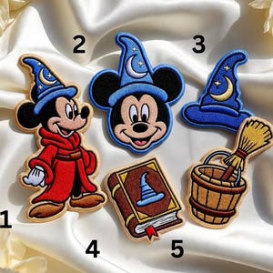 May include: Embroidered patches featuring a cartoon mouse in a wizard's hat and robe, along with a wizard hat, a book, and a broom. The patches are in shades of blue, red, brown, and yellow, with black and white accents. The patches are on a white satin surface.