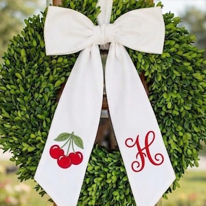 May include: A circular green boxwood wreath adorned with a large white fabric bow. The left tail of the bow features a graphic of two red cherries with green stems, while the right tail displays the red cursive letter H in a decorative font.