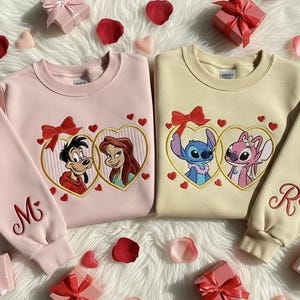 May include: Two sweatshirts, one pink and one beige, with embroidered cartoon characters in heart-shaped frames. The pink sweatshirt has the letter "M" embroidered on the sleeve. The beige sweatshirt has the letter "R" embroidered on the sleeve. The sweatshirts are surrounded by gift boxes and rose petals.