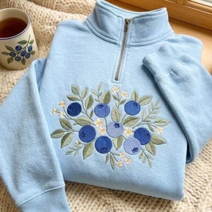 Op de afbeelding: Een lichtblauw sweatshirt met een korte rits en een geborduurd ontwerp van bosbessen, groene bladeren en kleine witte bloemetjes op de voorzijde. Een keramische mok met een bijpassend bosbessenpatroon en een mandje met bosbessen staan ernaast.