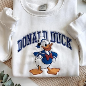May include: White sweatshirt featuring a graphic of Donald Duck in his sailor outfit, with the text "DONALD DUCK" in blue arched lettering. The sweatshirt has a crew neck and long sleeves.