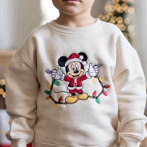 Puede incluir: Sudadera color crema con un diseño festivo de Mickey Mouse. Mickey viste un traje de Papá Noel y sostiene una guirnalda de luces navideñas de colores. La sudadera es una prenda cómoda de manga larga, perfecta para la temporada navideña.