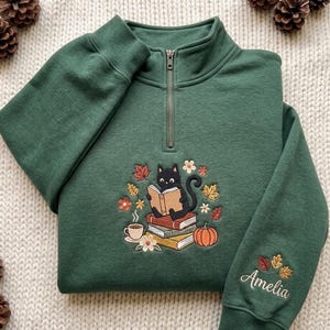 Puede incluir: Una sudadera verde oscuro con cremallera corta y un bordado de un gato negro leyendo sobre una pila de libros, rodeado de hojas de otoño, una calabaza y una taza de café. El nombre Amelia está bordado en la manga de la prenda.