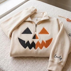 May include: A cream-colored quarter-zip sweatshirt featuring a large embroidered jack-o'-lantern face on the front. The face is split into black and orange sections. The name Emily is embroidered in black cursive script on the left sleeve cuff.