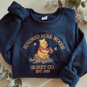 Puede incluir: Sudadera azul marino con un dise&ntilde;o bordado. Presenta a Winnie the Pooh sosteniendo un tarro de miel, con el texto "Hundred Acre Woods Honey Co. Est. 1926". El dise&ntilde;o incluye abejas y un toc&oacute;n de &aacute;rbol.