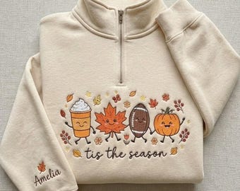 Puede incluir: Una sudadera color crema con cremallera corta y bordados de personajes otoñales, incluyendo una taza de café, una hoja de arce, un balón de fútbol americano y una calabaza. El texto 'tis the season' está bordado debajo y el nombre 'Amelia' en la manga.