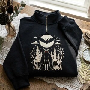 May include: A black quarter-zip sweatshirt featuring a cream-colored graphic print of a moth with red eyes standing in a forest with pine trees and a tree stump, beneath a large full moon and a flying bat surrounded by small stars and clouds.