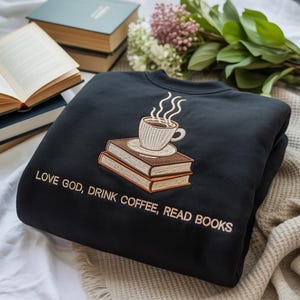 May include: Black sweatshirt featuring an embroidered coffee cup and books design, with the text "LOVE GOD, DRINK COFFEE, READ BOOKS". The sweatshirt is folded and placed on a white surface, with books and flowers in the background.