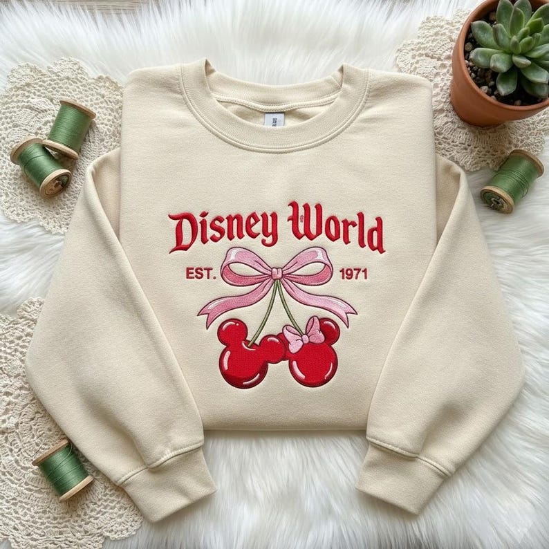 May include: Cream-colored sweatshirt with the words "Disney World" in red script, along with "EST. 1971." A pink bow and Mickey Mouse-shaped cherries are below the text. The sweatshirt is on a white fluffy surface.