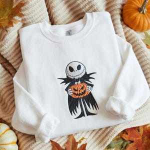 May include: A white long-sleeve sweatshirt featuring an embroidered graphic of a skeletal character with a round white head, black eye sockets, and a stitched mouth, wearing a black suit and holding a bright orange carved pumpkin.