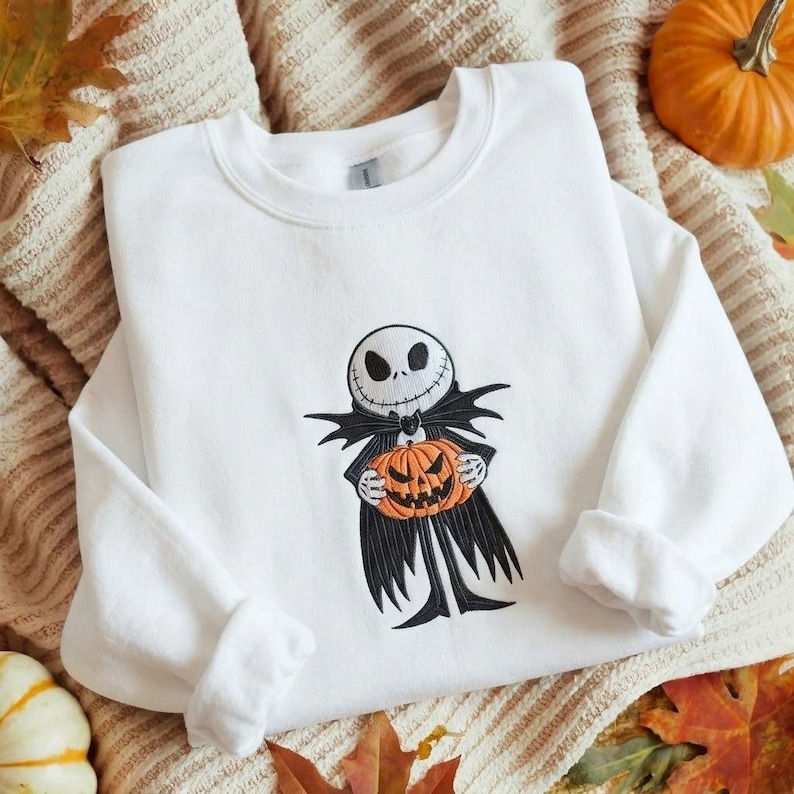 May include: A white long-sleeve sweatshirt featuring an embroidered graphic of a skeletal character with a round white head, black eye sockets, and a stitched mouth, wearing a black suit and holding a bright orange carved pumpkin.