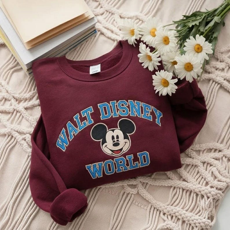 Puede incluir: Una sudadera de cuello redondo color granate con un gr&aacute;fico de Mickey Mouse centrado entre el texto arqueado "WALT DISNEY" y la palabra "WORLD" debajo. Las letras son de color azul claro con un borde blanco y tiene mangas largas.