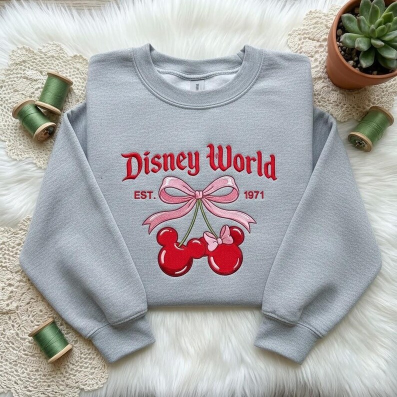 May include: A light gray sweatshirt with the words "Disney World" embroidered in red script. Below the text is a pink bow and two red cherry-shaped Mickey Mouse heads. The year "1971" is also embroidered on the sweatshirt.