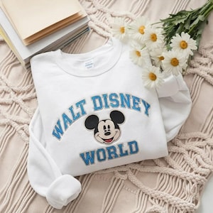 Puede incluir: Una sudadera blanca de cuello redondo con una cabeza de Mickey Mouse bordada en el centro, rodeada por el texto azul Walt Disney World. El texto est&aacute; dispuesto en arco sobre el personaje y en l&iacute;nea recta debajo del personaje.
