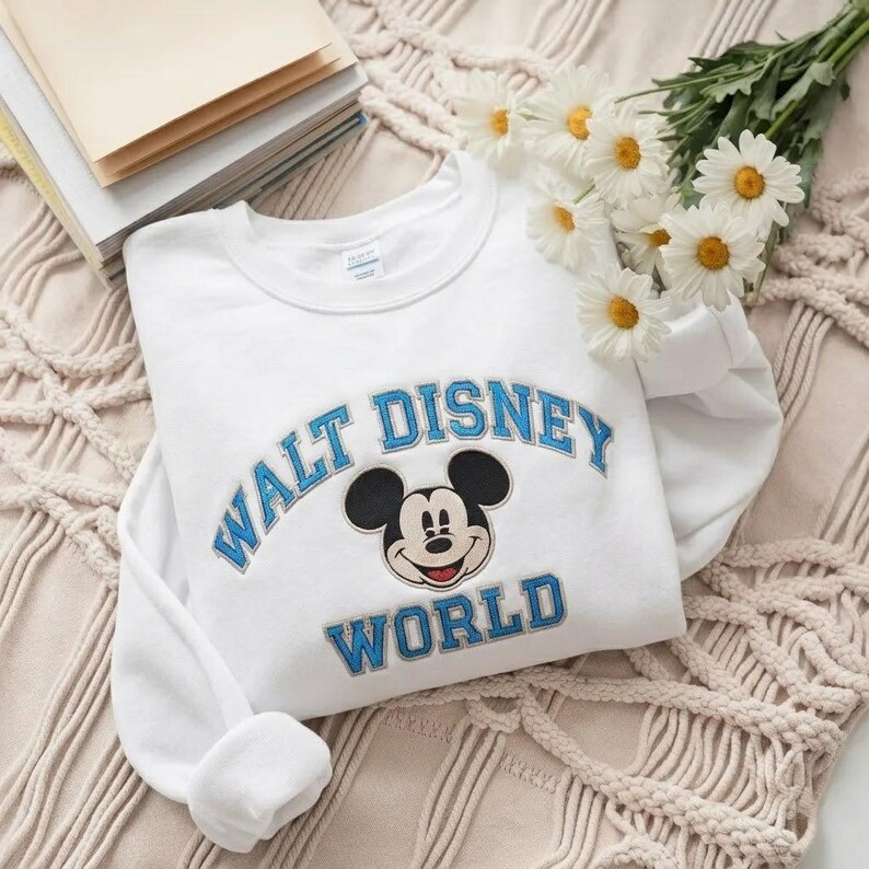 Puede incluir: Una sudadera blanca de cuello redondo con una cabeza de Mickey Mouse bordada en el centro, rodeada por el texto azul Walt Disney World. El texto est&aacute; dispuesto en arco sobre el personaje y en l&iacute;nea recta debajo del personaje.