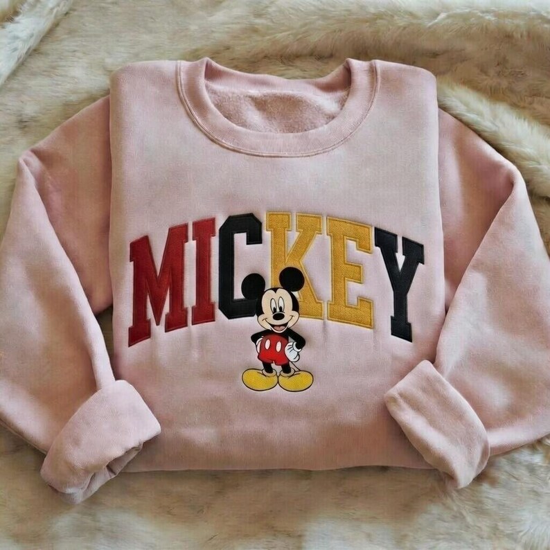 Puede incluir: Una sudadera rosa claro con la palabra "MICKEY" en letras de bloque rojas, negras y amarillas. Una imagen de dibujos animados de Mickey Mouse est&aacute; centrada debajo del texto. La sudadera tiene cuello redondo y mangas largas.