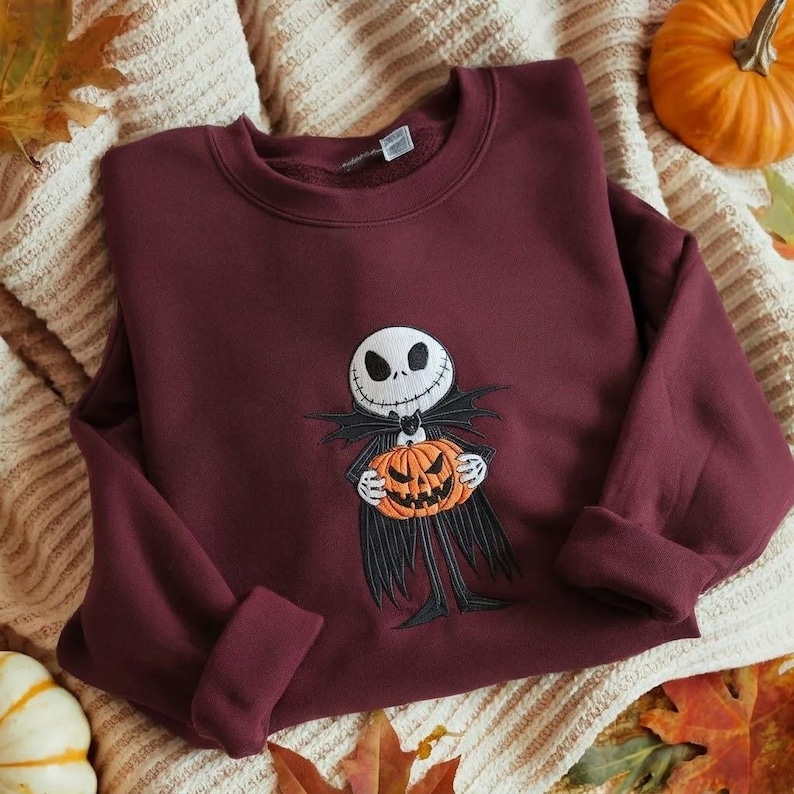 May include: A maroon long-sleeve sweatshirt featuring a central embroidered graphic of a skeletal character with a white skull face, wearing a black suit and holding a bright orange carved jack-o'-lantern pumpkin with a jagged mouth.