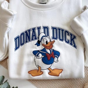 May include: White sweatshirt featuring a graphic of Donald Duck in a sailor outfit, with the text "DONALD DUCK" in blue arched lettering. The sweatshirt has long sleeves and a crew neck.