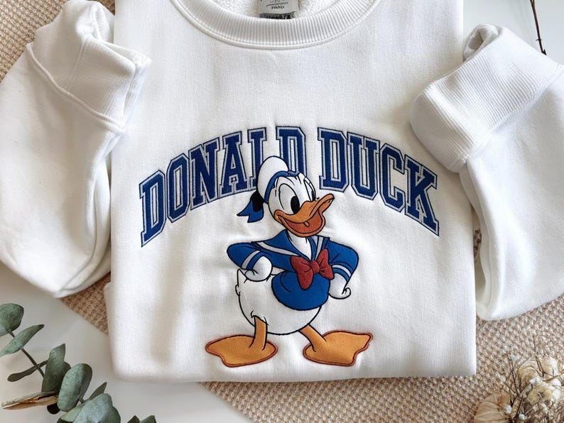 May include: White sweatshirt featuring a graphic of Donald Duck in a sailor outfit, with the text "DONALD DUCK" in blue arched lettering. The sweatshirt has long sleeves and a crew neck.