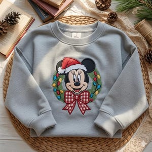 May include: A light gray sweatshirt featuring a festive Minnie Mouse design. Minnie is wearing a Santa hat and is surrounded by a wreath with colorful lights and a red and white checkered bow. The sweatshirt has long sleeves.