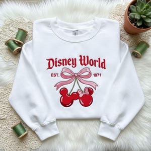 May include: White crewneck sweatshirt with a red "Disney World" design, featuring a bow and Mickey Mouse-shaped cherries. The text "EST. 1971" is also included. The sweatshirt is a casual style.