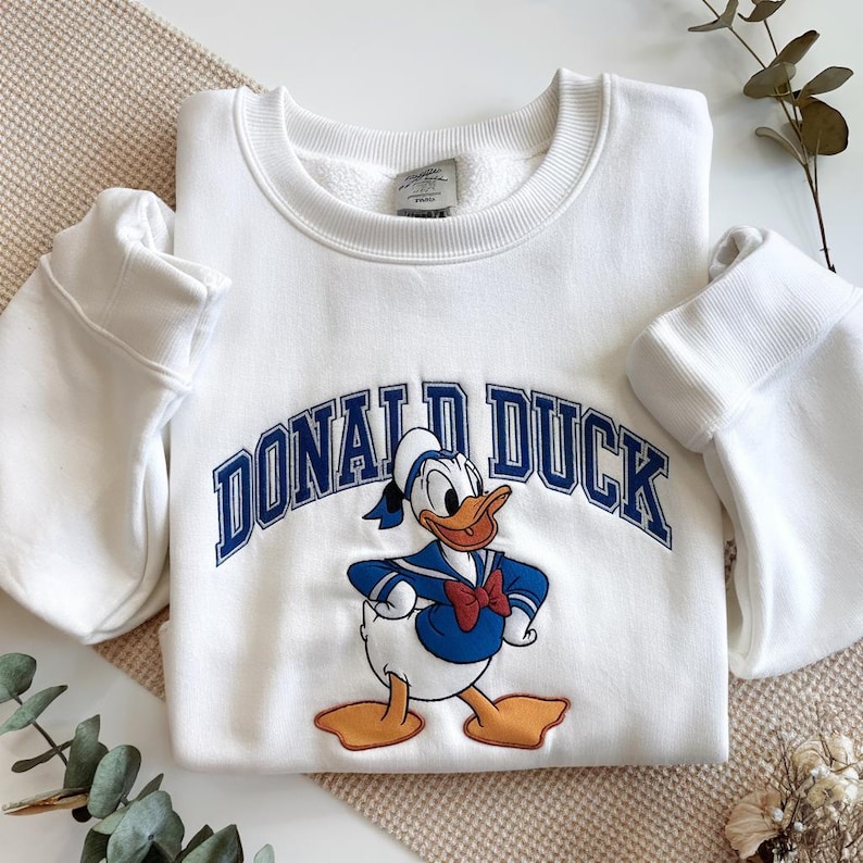 May include: White sweatshirt featuring a graphic of Donald Duck in his sailor outfit, with the text "DONALD DUCK" in blue arched lettering. The sweatshirt has a crew neck and long sleeves.