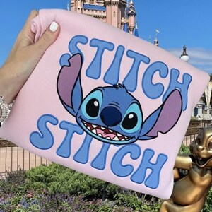 Puede incluir: Una sudadera rosa claro con el personaje Stitch de la pel&iacute;cula Lilo & Stitch. La palabra "Stitch" est&aacute; escrita en azul, con un gr&aacute;fico azul de Stitch. El fondo incluye un castillo y una estatua.